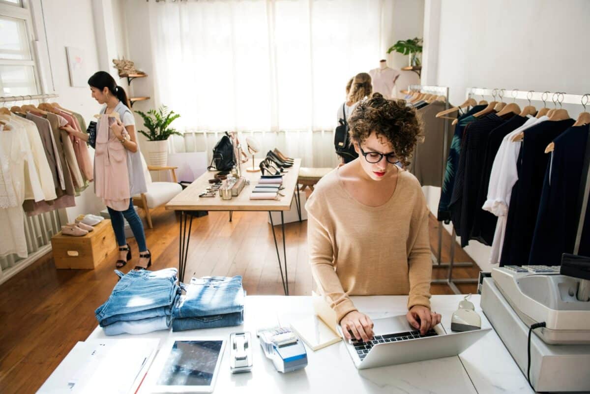 Woman working at a laptop in a clothing store, surrounded by racks of clothes and stacks of jeans.