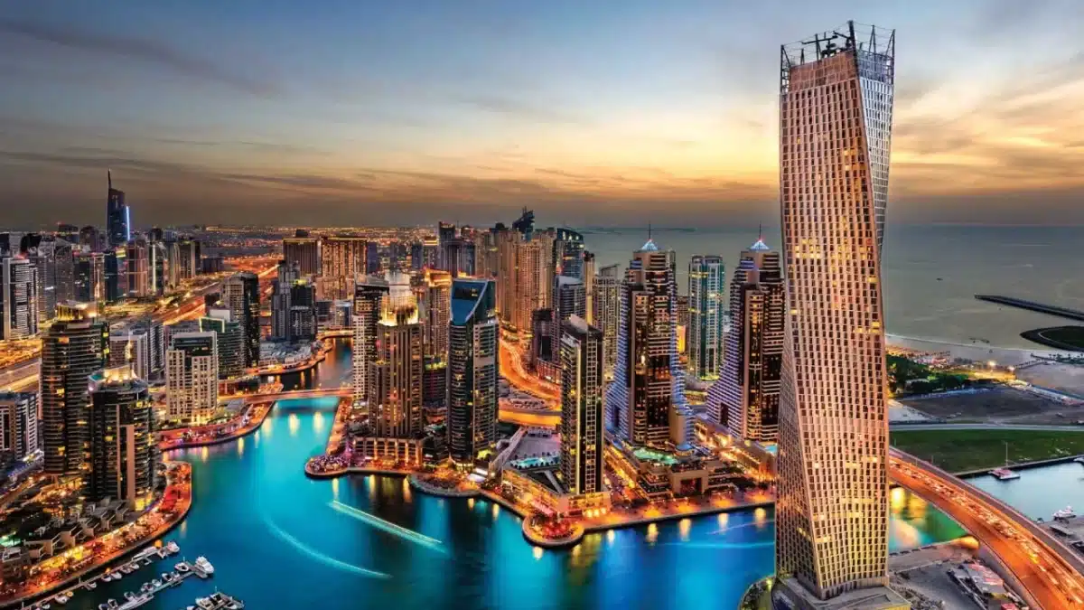 Ras Al Khaimah Free Trade Zone company setup A stunning aerial view of a cityscape at dusk, showing tall, brightly lit skyscrapers surrounding a winding body of water. The sky glows with warm hues of a setting sun, and the city lights reflect off the water, creating a vibrant and dynamic scene—perfect for establishing a company in the Ras Al Khaimah Free Trade Zone.