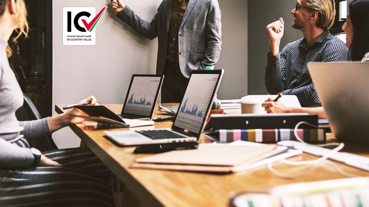 A group of four people in a modern office setting, seated around a wooden table with laptops, documents, and coffee. One person stands and presents, gesturing at a screen. The ICV Services Dubai logo is visible on the wall.